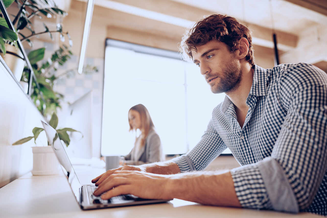 Jovem trabalha em notebook em um escritório com colega ao fundo 