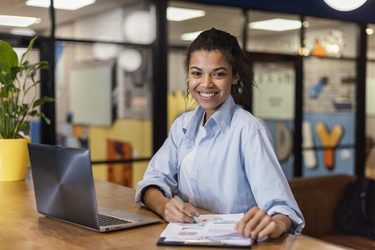 Jovem assitente administrativa posa sorridente enquanto trabalha em seu notebook 